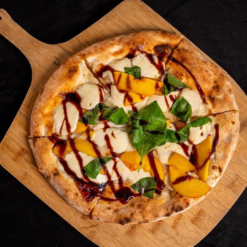 Round artisan pizza topped with mozzarella, peach slices, fresh basil, and balsamic glaze on a wooden serving board.