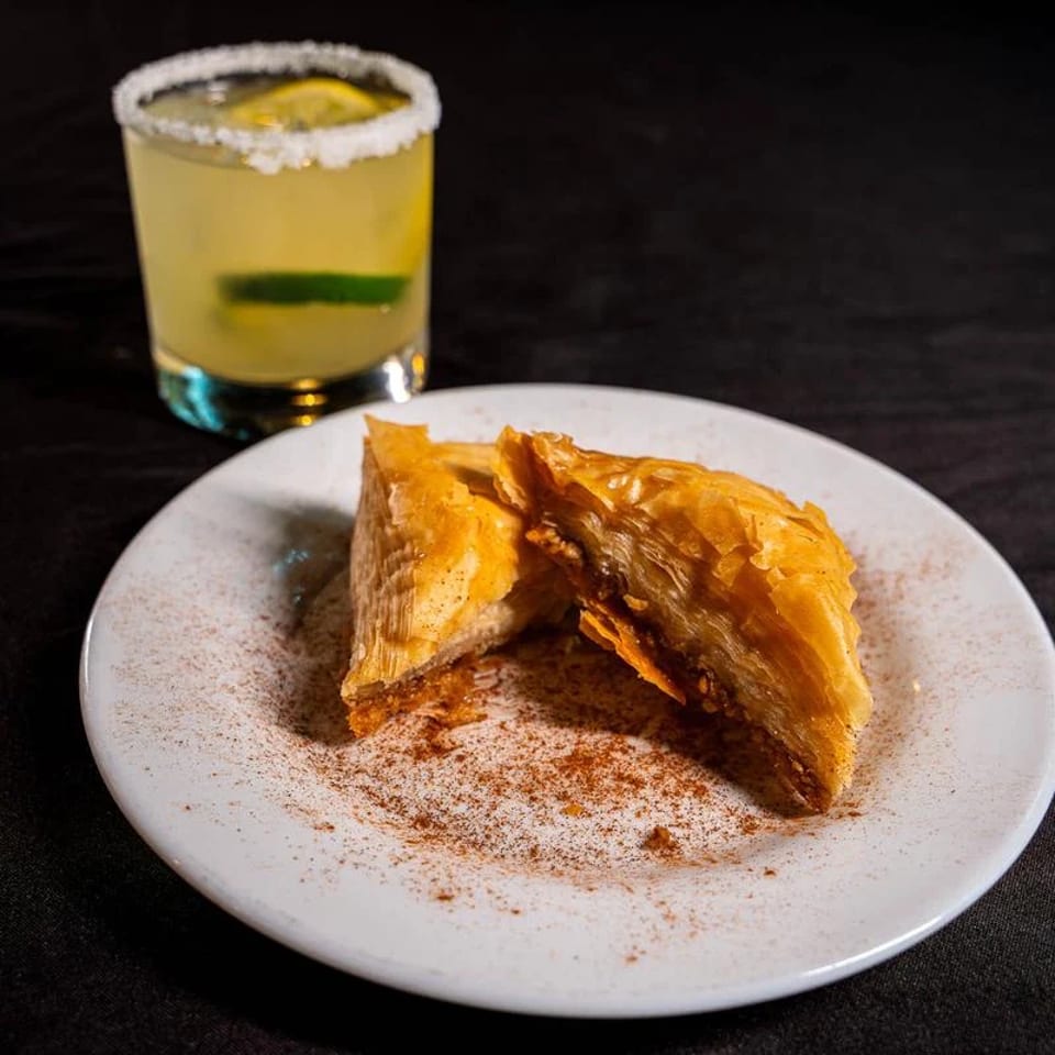 Two golden baklava pastries sit on a cinnamon-dusted plate beside a yellow cocktail with a salted rim and lime garnish.