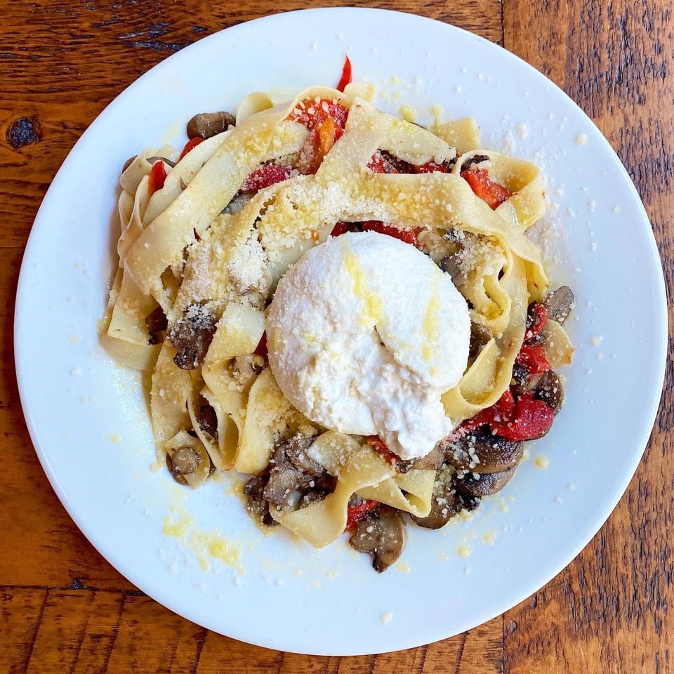 Wide ribbon pasta topped with mushrooms, roasted tomatoes, grated cheese, and a dollop of creamy ricotta on a white plate.