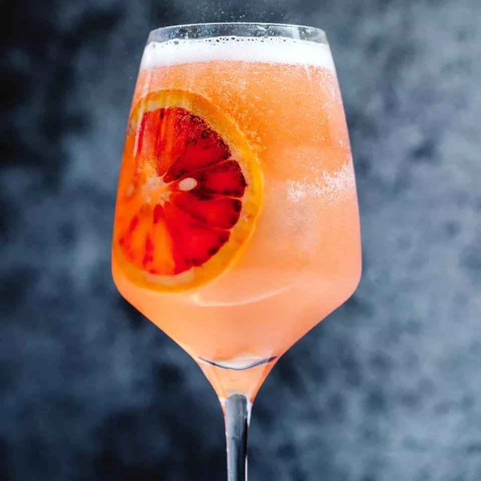 Orange cocktail with a frothy top and blood orange slice garnish in a stemmed glass against a smoky dark background.