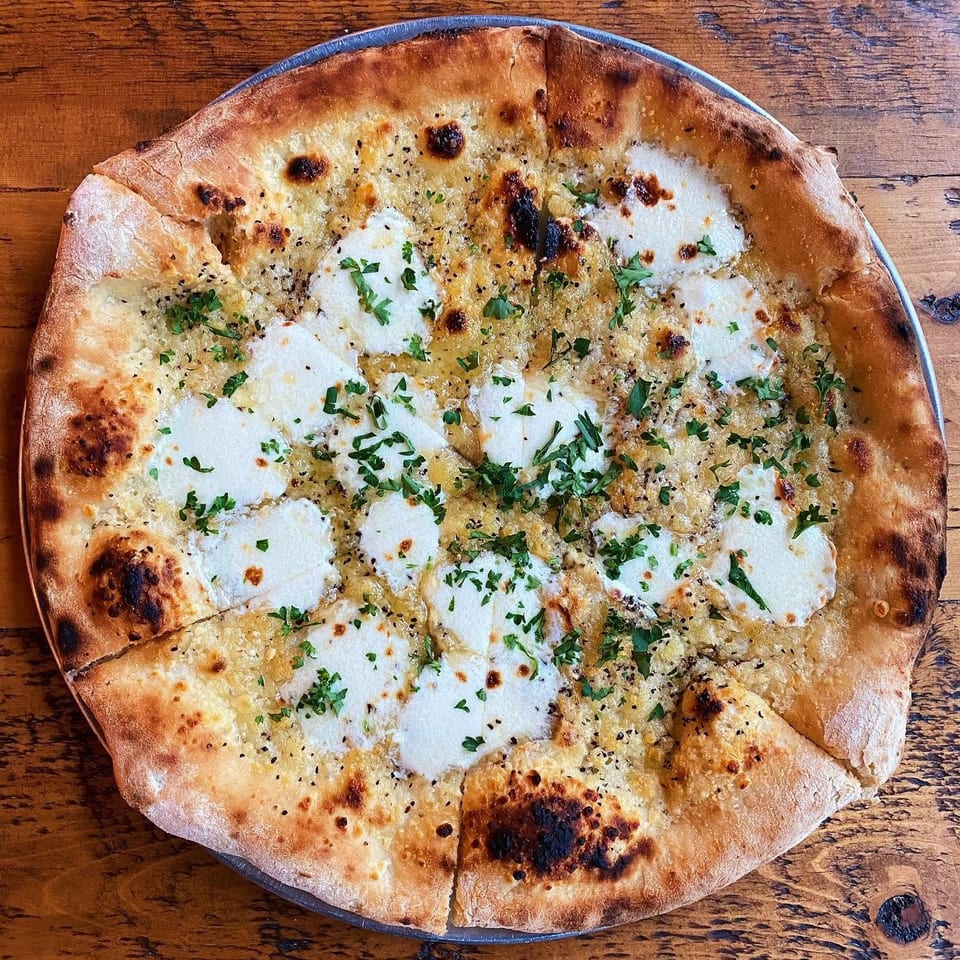 Overhead view of a blistered flatbread topped with melted white cheese, garlic, herbs, and black pepper on a wooden table.