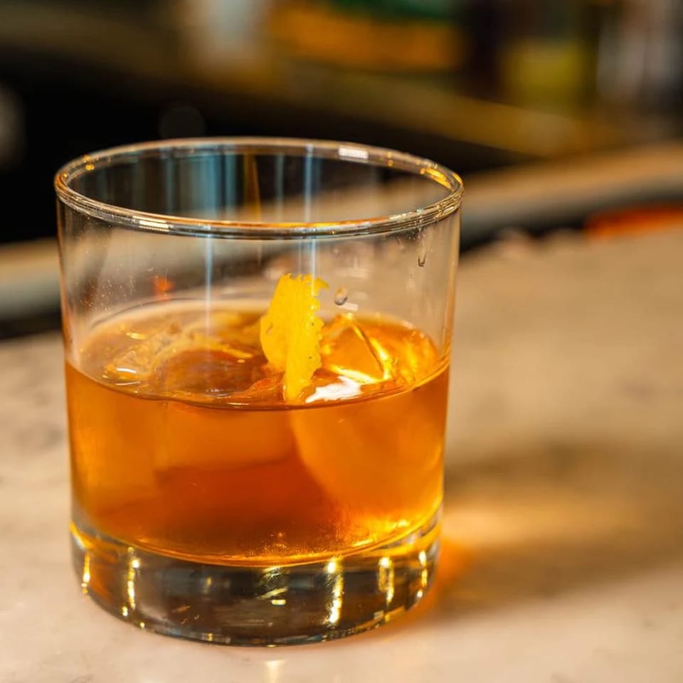 Amber cocktail with a citrus peel garnish served over ice in a rocks glass on a warmly lit bar.