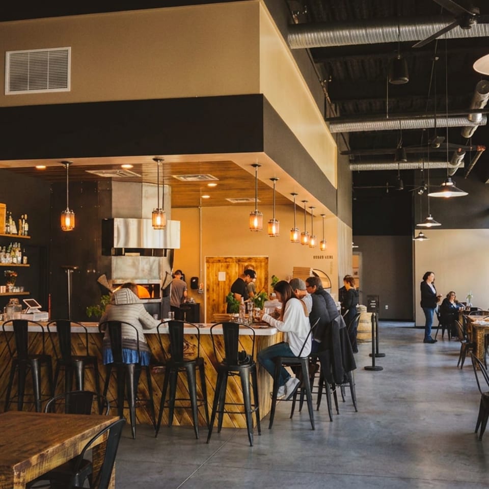 Modern restaurant interior with a long bar, pendant lighting, exposed ductwork, wooden tables, and diners seated near large windows.