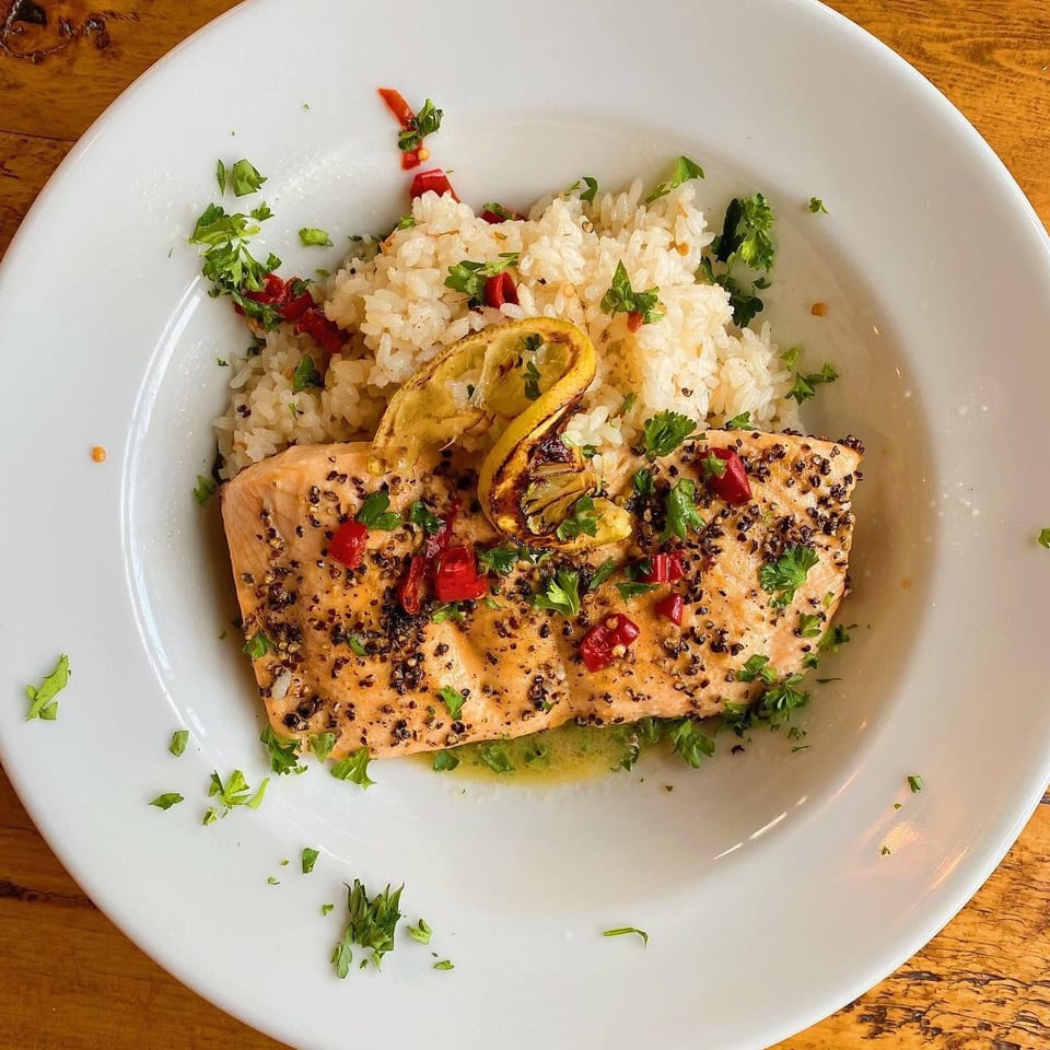 Grilled pepper-crusted salmon with rice, charred lemon, red chilies, and parsley on a white plate.