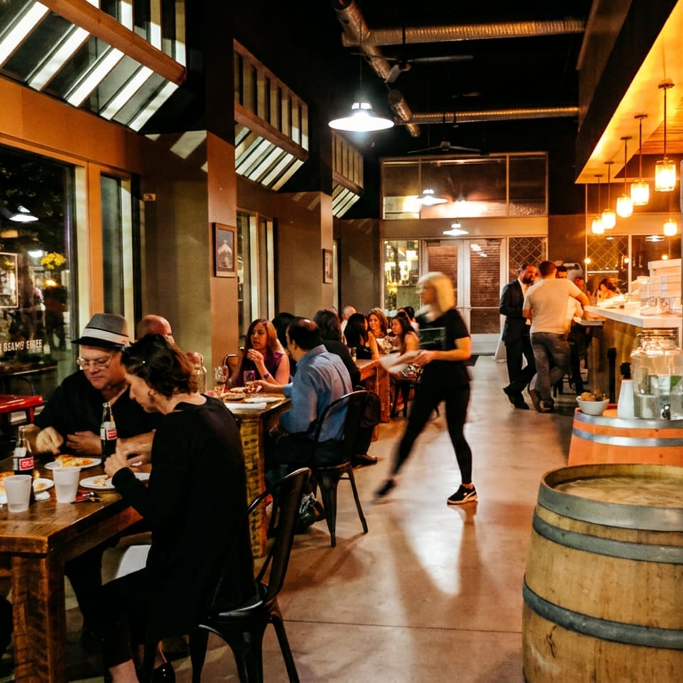 Busy warmly lit restaurant dining room with patrons eating, servers walking, exposed beams, skylights, and wooden barrels near the bar.