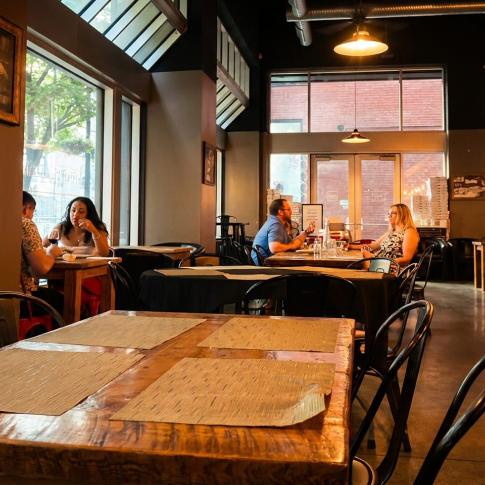 Warmly lit restaurant dining room with wooden tables, black chairs, exposed ductwork, large windows, and several diners.