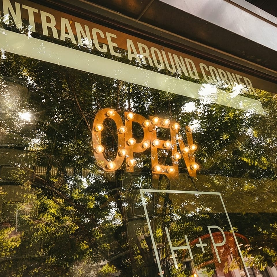 Restaurant storefront window displaying a glowing OPEN sign, entrance-around-corner notice, and leafy tree reflections.